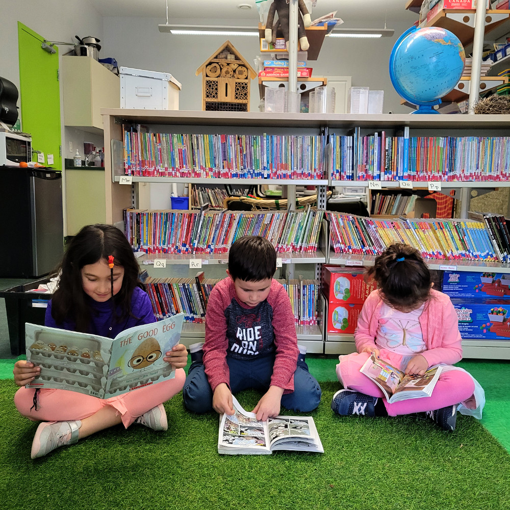 three children sitting in a row reading infront of a bookshelf full of books